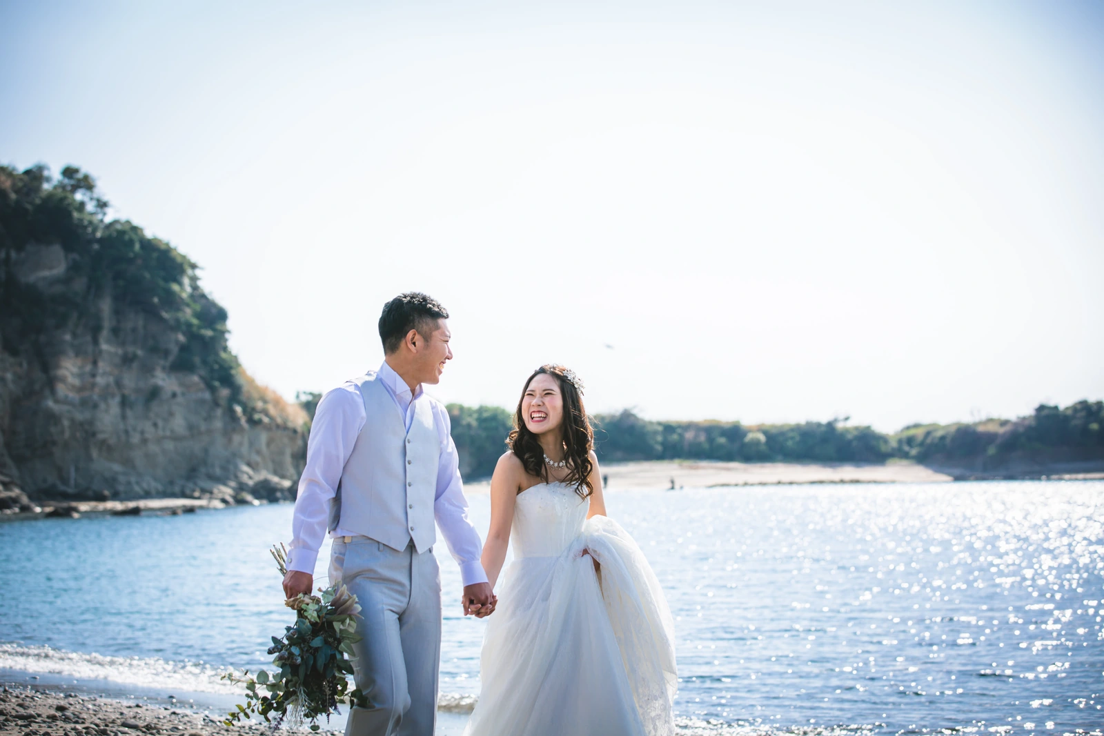 長者ヶ崎海岸の渚を手をつないで歩くウェディングドレスの花嫁とタキシードの新郎