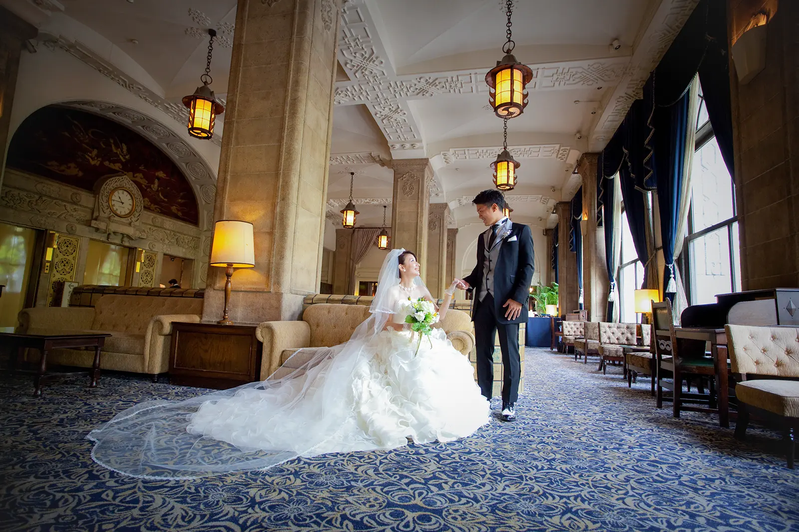 クラシカルなロビー空間で新郎が立ち新婦が座って見つめ合うフォトウェディング写真