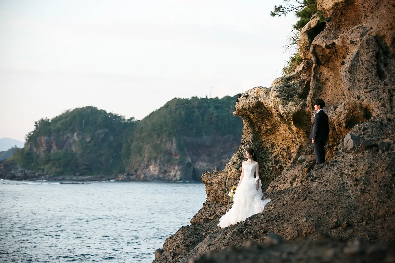 巌門の断崖と入り江を背景にウェディングドレスで岩場に立つ花嫁と新郎