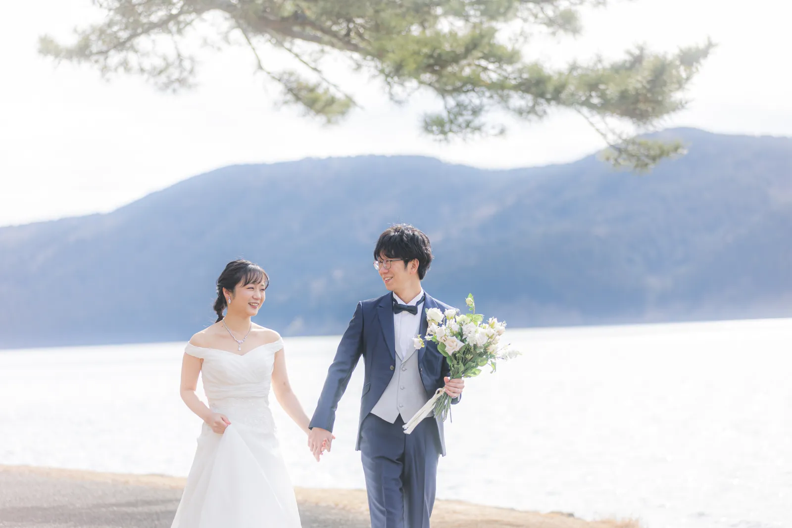 冬の芦ノ湖畔で光を背に受けて自然に微笑む洋装の新郎新婦