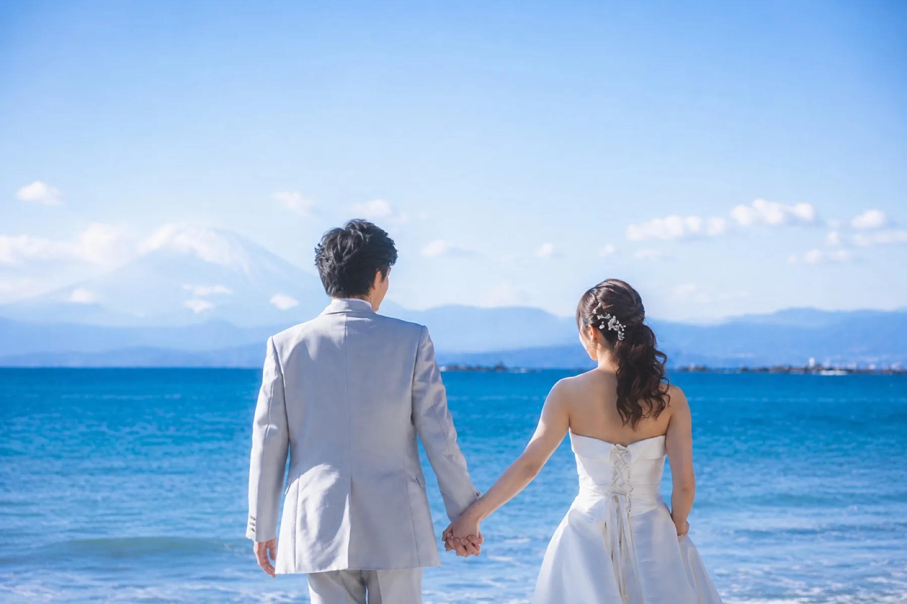 葉山の海辺で手をつなぎ富士山を望む新郎新婦、洋装フォトウェディングのロケーション撮影