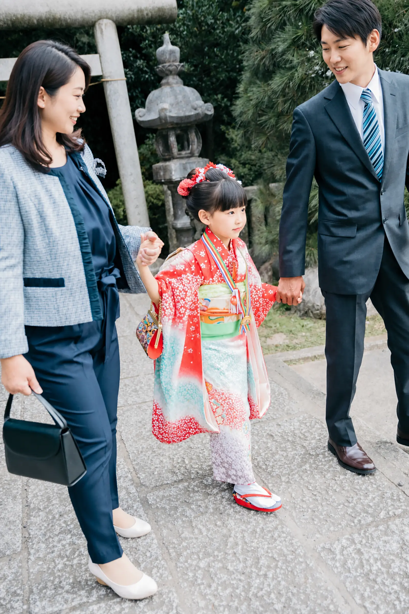 石灯籠と鳥居のそばで両手をご家族とつないで歩く7歳の女の子、四つ身の七五三（本牧神社・横浜）