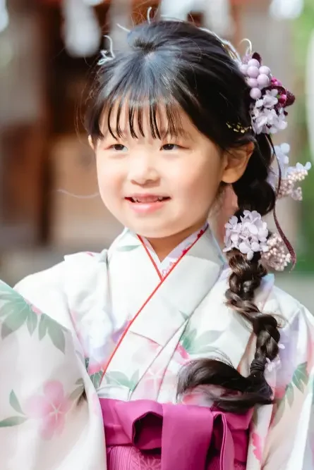千歳飴の箱を手に、はにかんで笑う7歳の女の子の上半身。太い三つ編みと藤色の髪飾りが見える七五三の一枚（星川杉山神社・横浜）