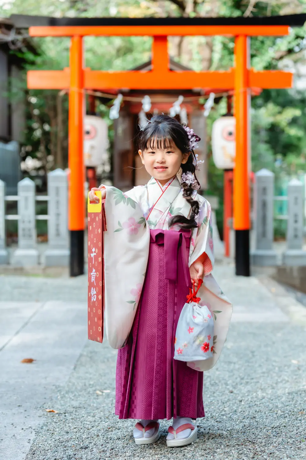 千歳飴と書かれた赤い箱を掲げて笑う、袴すがたの7歳の女の子。星川杉山神社の七五三出張撮影（横浜）