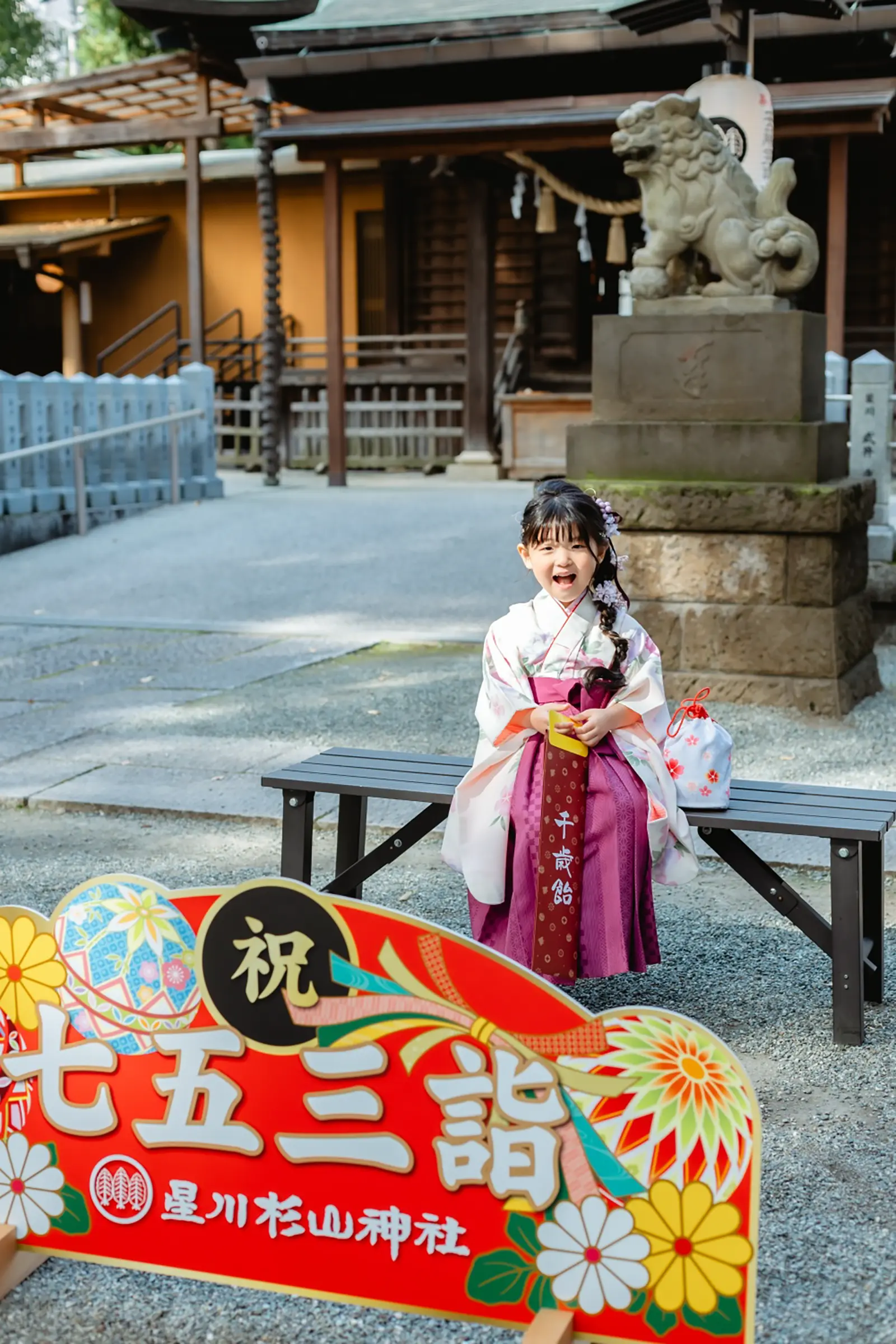 「祝 七五三詣 星川杉山神社」の装飾看板の奥、ベンチに腰かけて千歳飴を抱える袴すがたの7歳の女の子（横浜の七五三出張撮影）