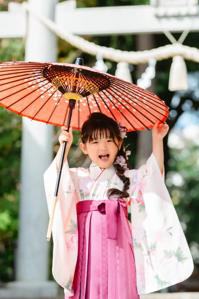赤い和傘を両手で頭上に掲げ、口を大きく開けて笑う袴すがたの7歳の女の子。星川杉山神社の七五三出張撮影（横浜）