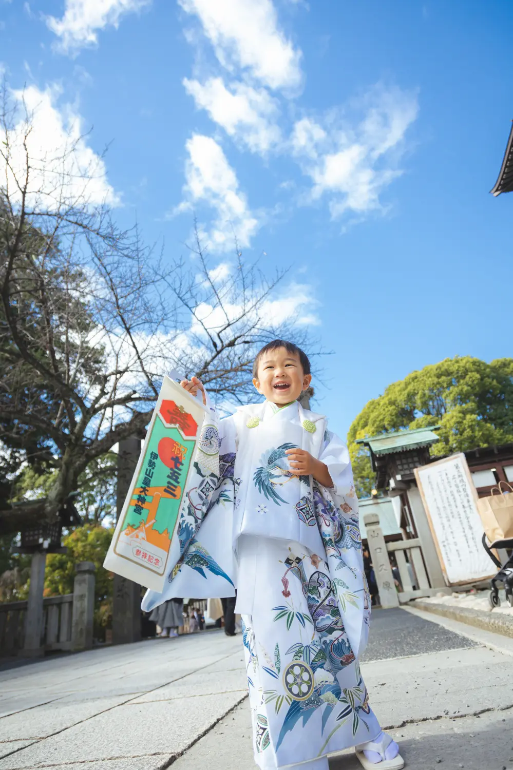 青空を背にローアングルで笑う3歳の男の子、白い被布に鷹柄の着物、伊勢山皇大神宮の七五三出張撮影（横浜）