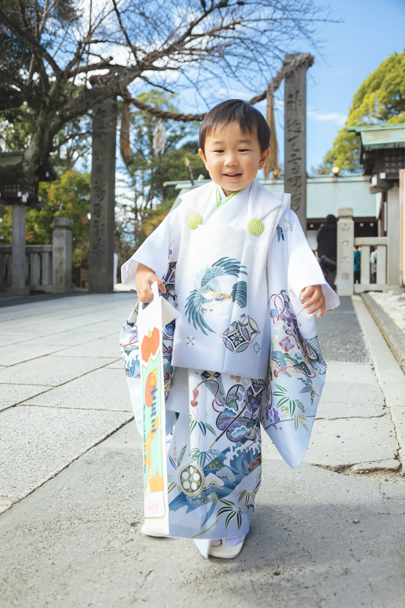 石鳥居の下を歩いてくる3歳の男の子の全身、白い被布に鷹柄の着物、伊勢山皇大神宮の七五三出張撮影（横浜）