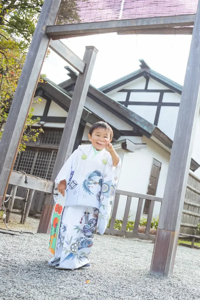 木の鳥居の下で顔に手を当てておどける3歳の男の子、白い被布姿の七五三（伊勢山皇大神宮・横浜）