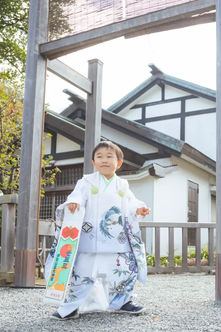 木の鳥居の下に立ち紙袋を提げた3歳の男の子の全身、白い被布姿の七五三（伊勢山皇大神宮・横浜）
