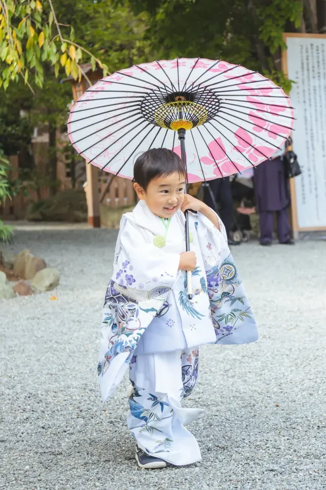 花模様の和傘をかついで玉砂利の上を歩く3歳の男の子の全身、白い被布姿の七五三（伊勢山皇大神宮・横浜）