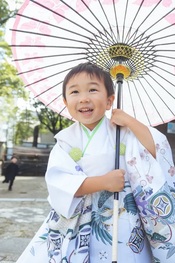 淡いピンクの花模様の和傘をさして笑う3歳の男の子、白い被布姿の七五三（伊勢山皇大神宮・横浜）