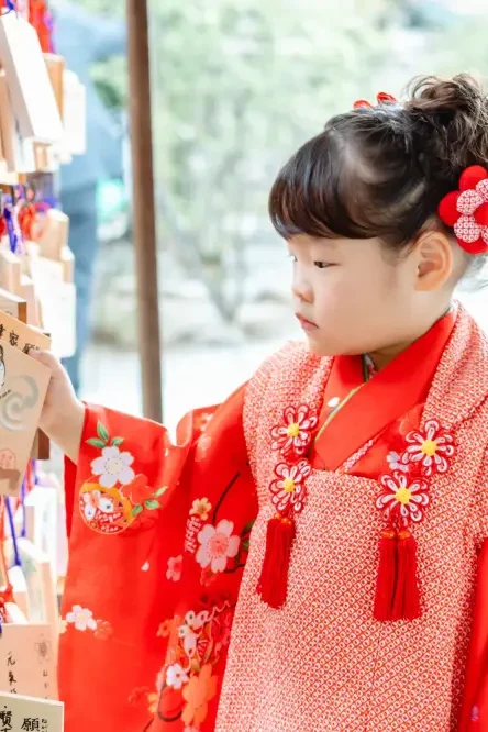 絵馬掛けの絵馬に手を伸ばす赤い被布姿の3歳の女の子（七五三・伊勢山皇大神宮）