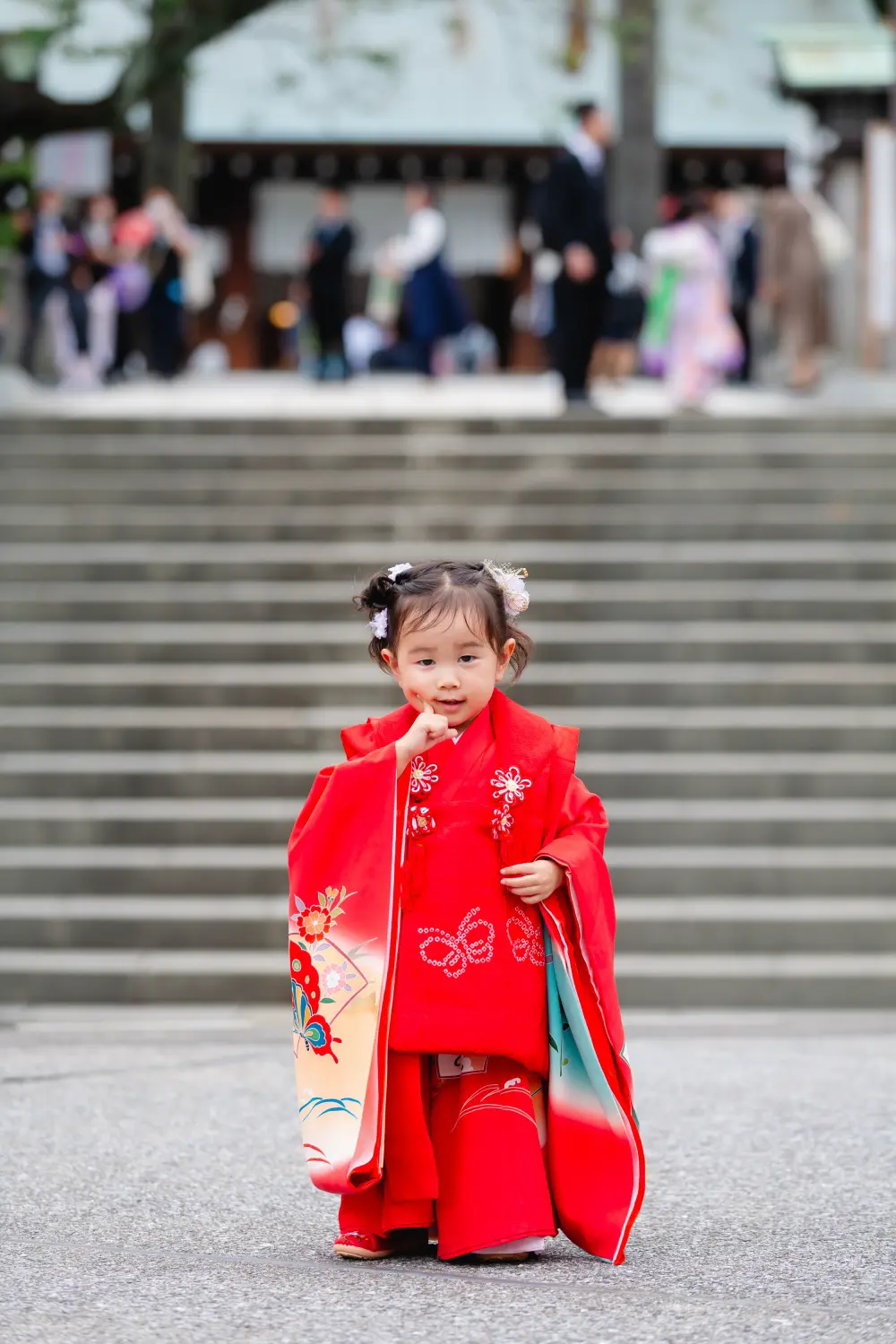 石段を背に指を口元にあてて立つ朱赤の被布姿の3歳の女の子、七五三（伊勢山皇大神宮・横浜）