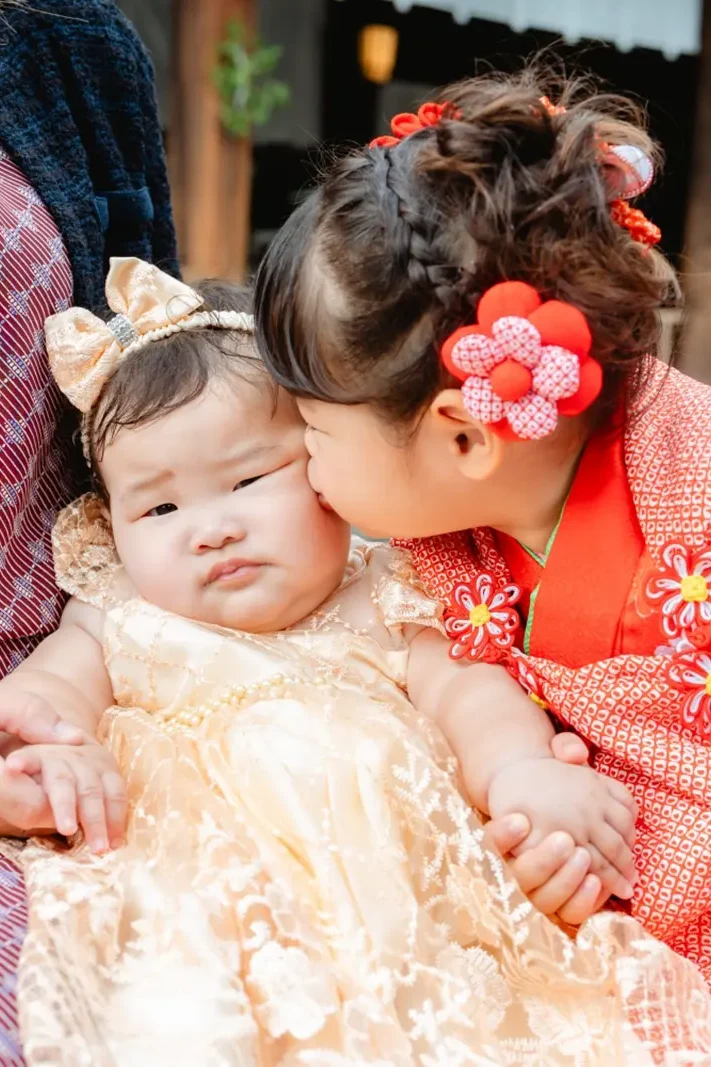 乳児の妹の頬に顔を寄せる、赤い被布姿の3歳の女の子（七五三・伊勢山皇大神宮）