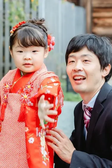 父としゃがんで並び笑う、赤い被布姿の3歳の女の子（七五三・伊勢山皇大神宮）