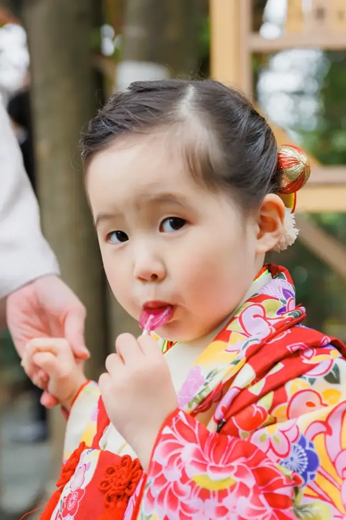 絵馬掛けを背景に棒つきの飴をくわえて横目でカメラを見る3歳の女の子、朱赤の被布（伊勢山皇大神宮・横浜の七五三）