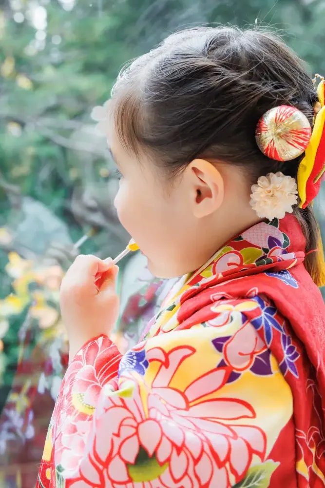 黄葉した木々のガラスの映り込みを背景に棒つきの飴を口に運ぶ3歳の女の子の横顔、朱赤の被布（伊勢山皇大神宮・横浜の七五三）