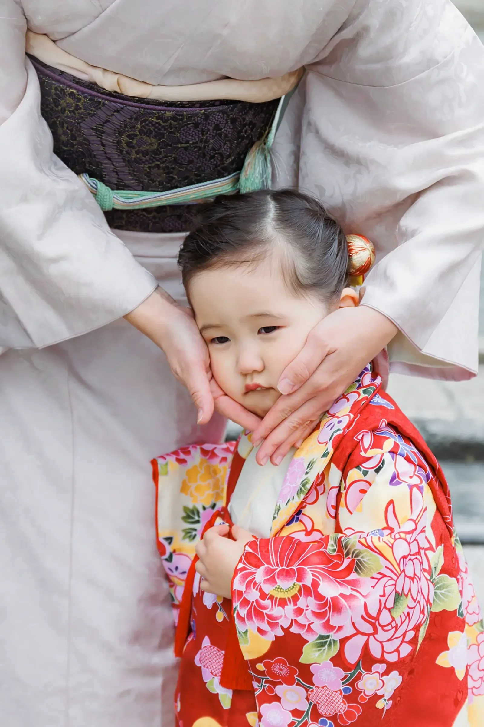母の着物と帯を背景に両頬を手で包まれ困った表情の3歳の女の子、朱赤の被布（伊勢山皇大神宮・横浜の七五三）