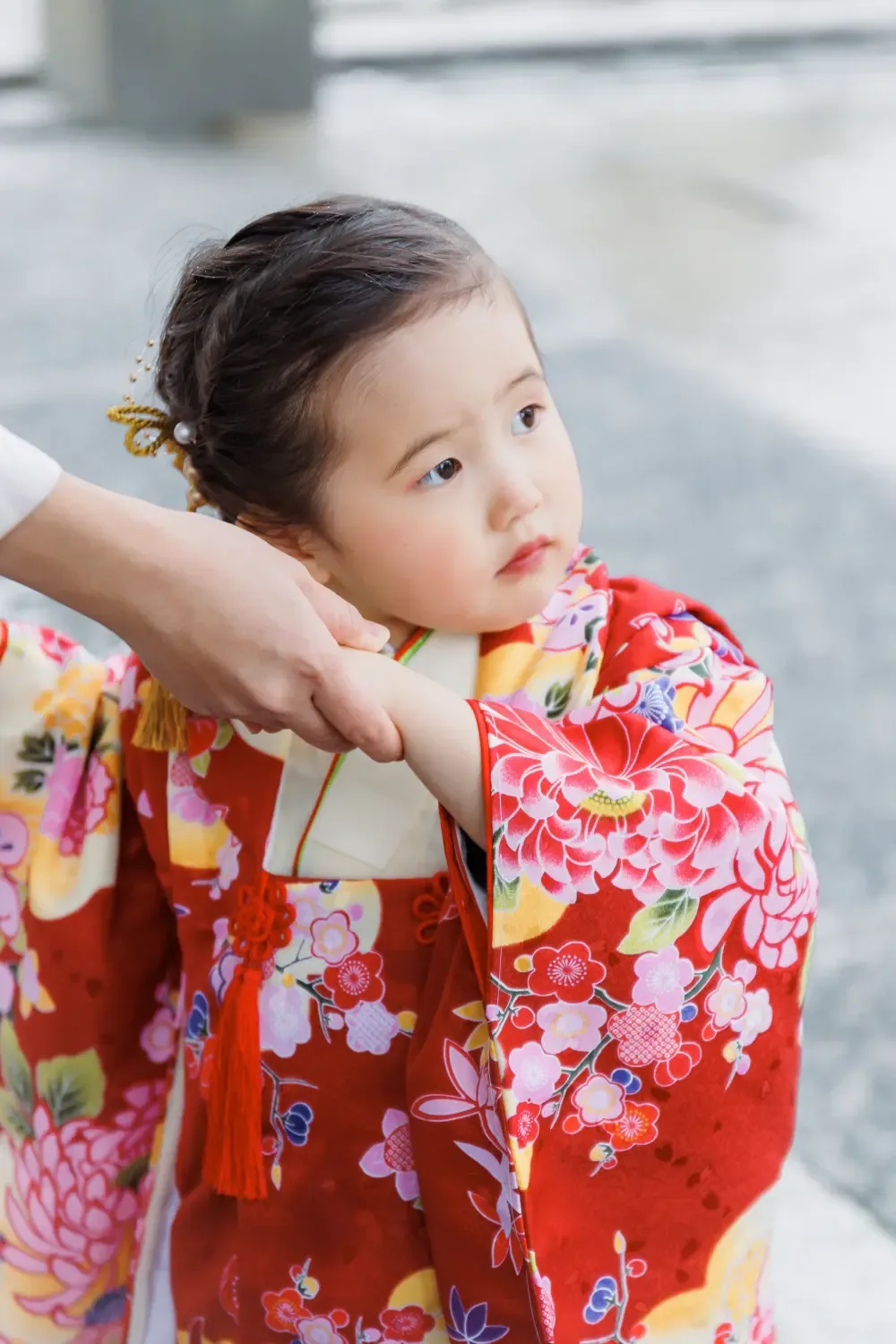 母に腕を取られカメラの外を見つめる3歳の女の子のバストアップ、朱赤の被布と玉かんざし（伊勢山皇大神宮・横浜の七五三）