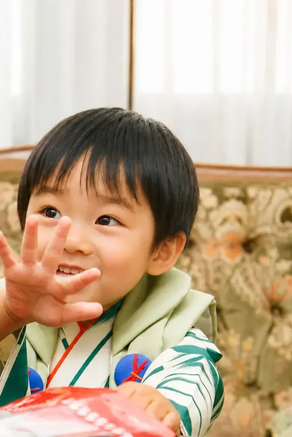 洋室のソファでお菓子に手を伸ばす3歳の男の子、深緑の着物と若草色の被布｜石浦神社の七五三出張撮影（金沢）