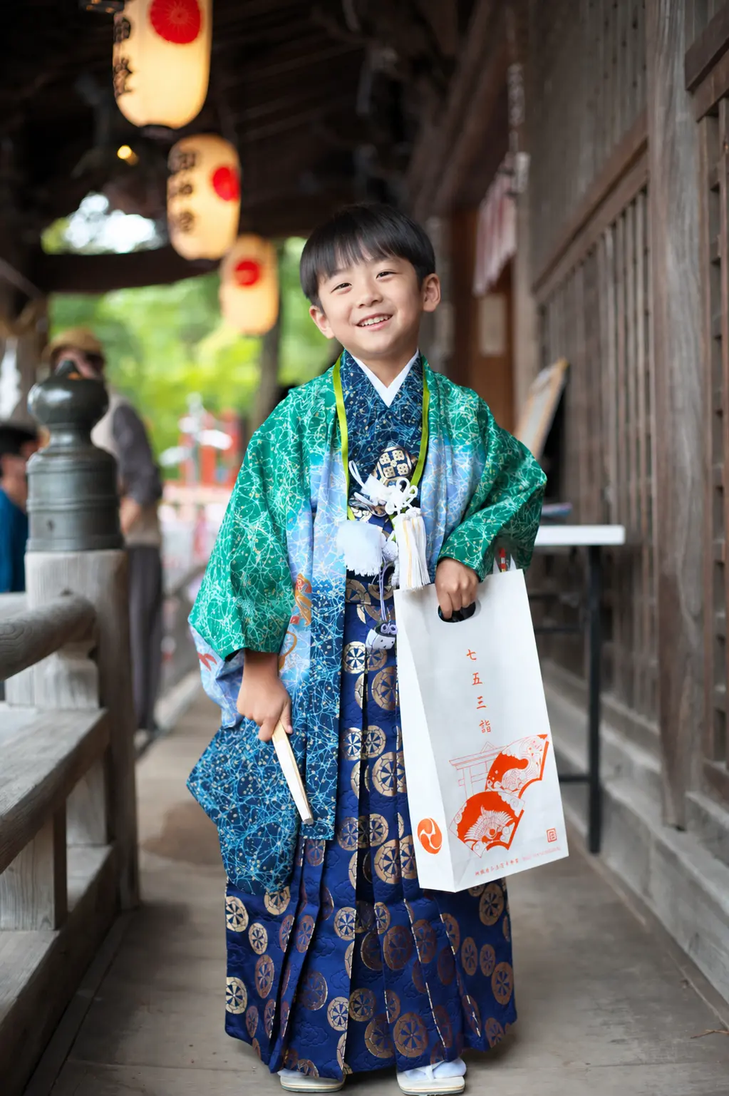 提灯が下がる回廊で授与品の袋を持って笑う5歳の男の子、緑の羽織袴の七五三（石浦神社・金沢）