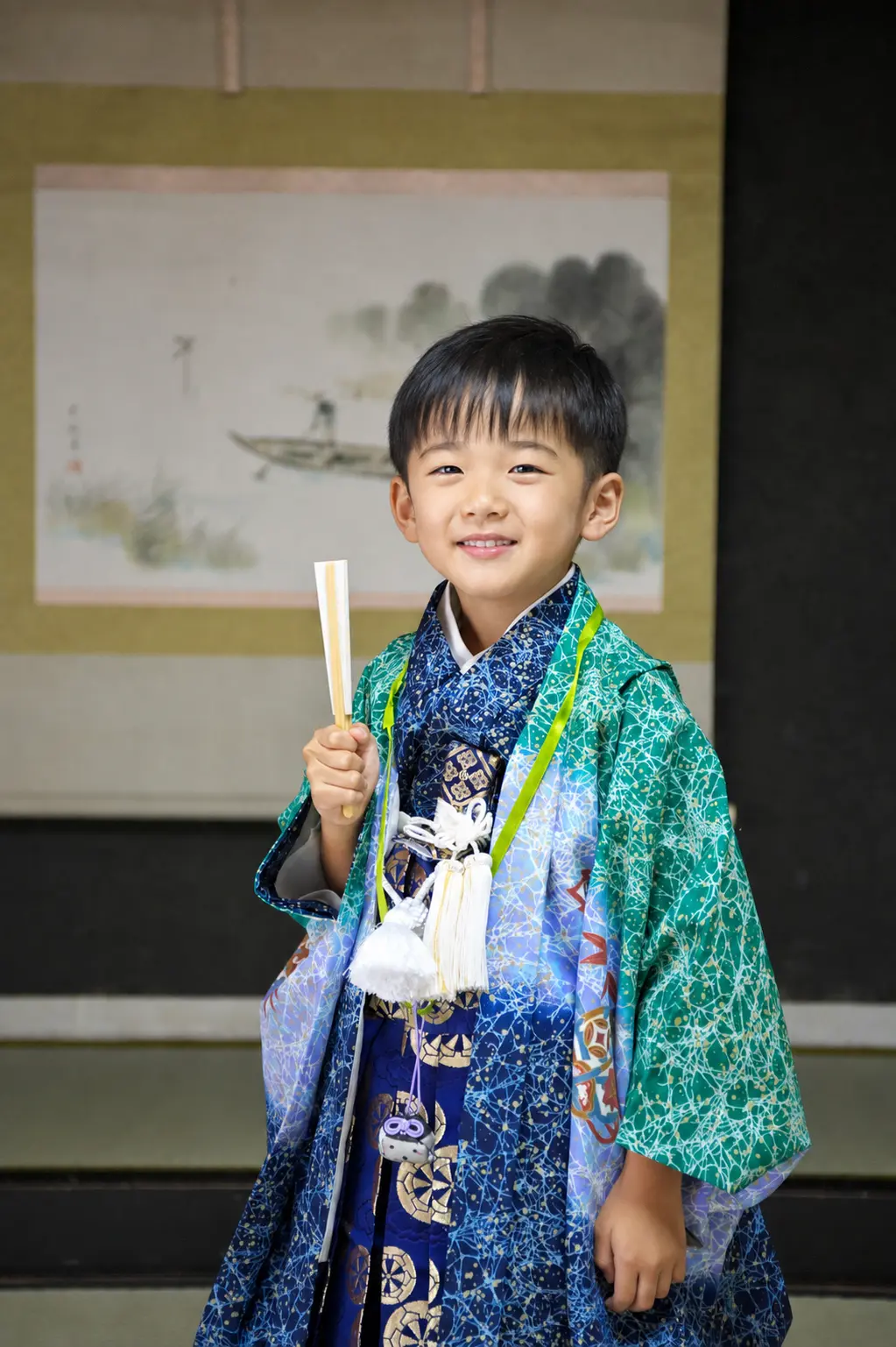 和室の掛軸前で細長い紙のものを手に微笑む5歳の男の子、緑の羽織袴の七五三