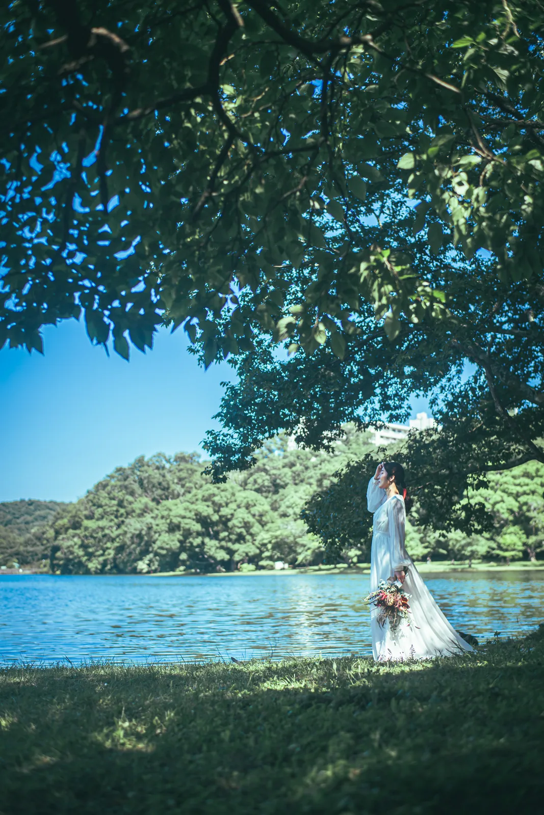 木陰から望む一碧湖の水辺でブーケを持つウェディングドレスの花嫁