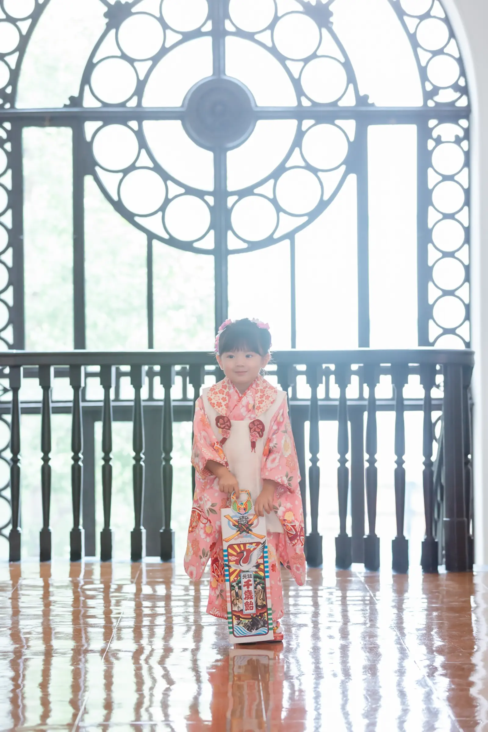 円形の鉄格子窓を背にして、細長い紙箱を両手で持ち微笑む被布姿の3歳の女の子の七五三出張撮影（横浜市開港記念会館）