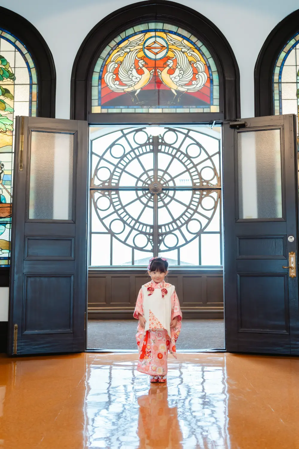 二羽の鳥が向かい合うステンドグラスの欄間と開いた扉の中央に立つ、被布姿の3歳の女の子の七五三出張撮影（横浜市開港記念会館）