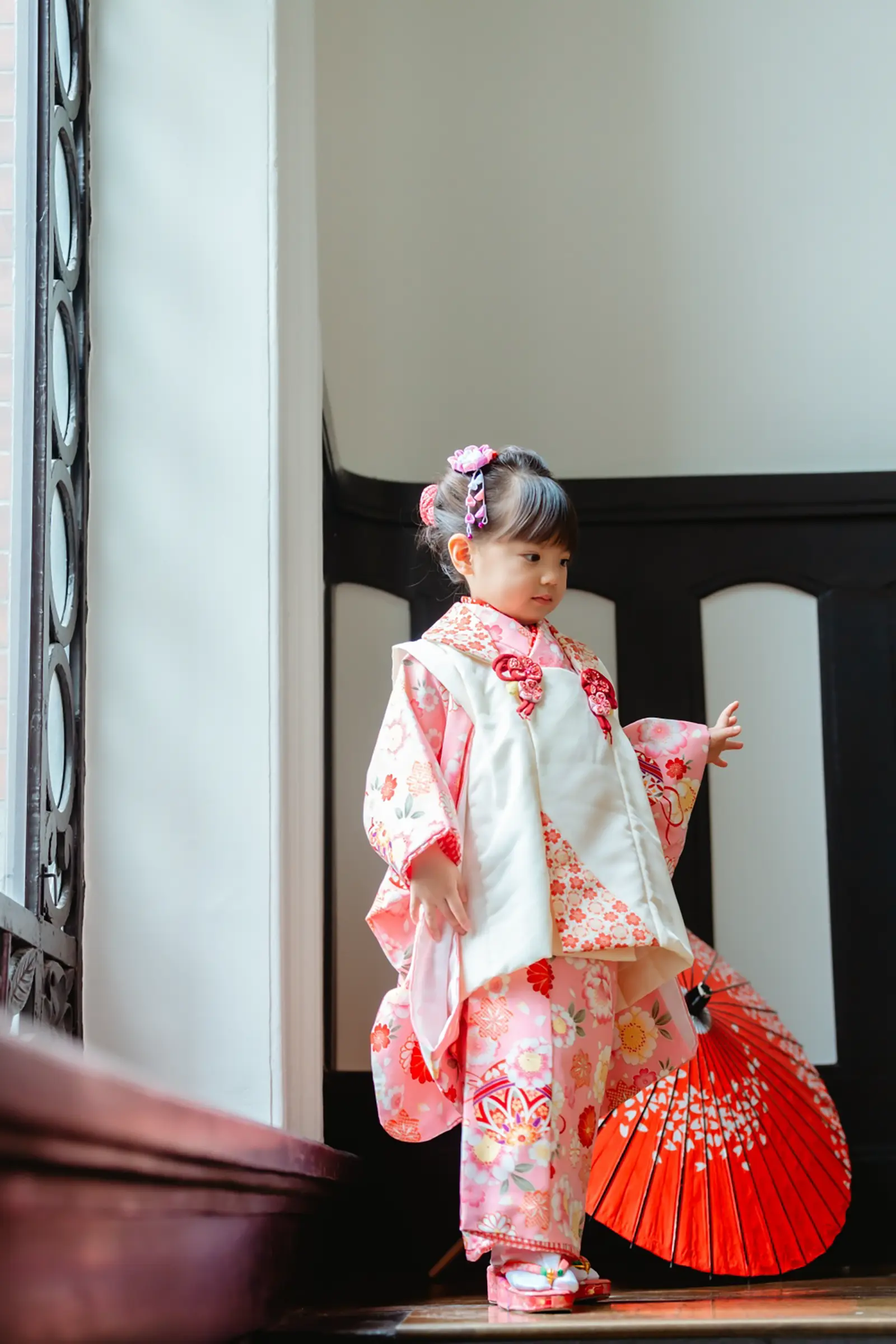 窓辺で朱色の和傘を引きずるように持って歩き出す、被布姿の3歳の女の子の七五三出張撮影（横浜市開港記念会館）
