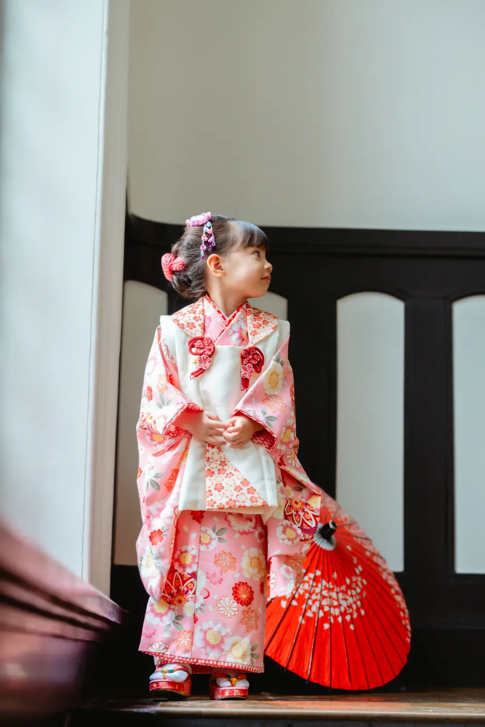 朱色の和傘を体の横に立たせ、横顔で微笑む被布姿の3歳の女の子の七五三出張撮影（横浜市開港記念会館）