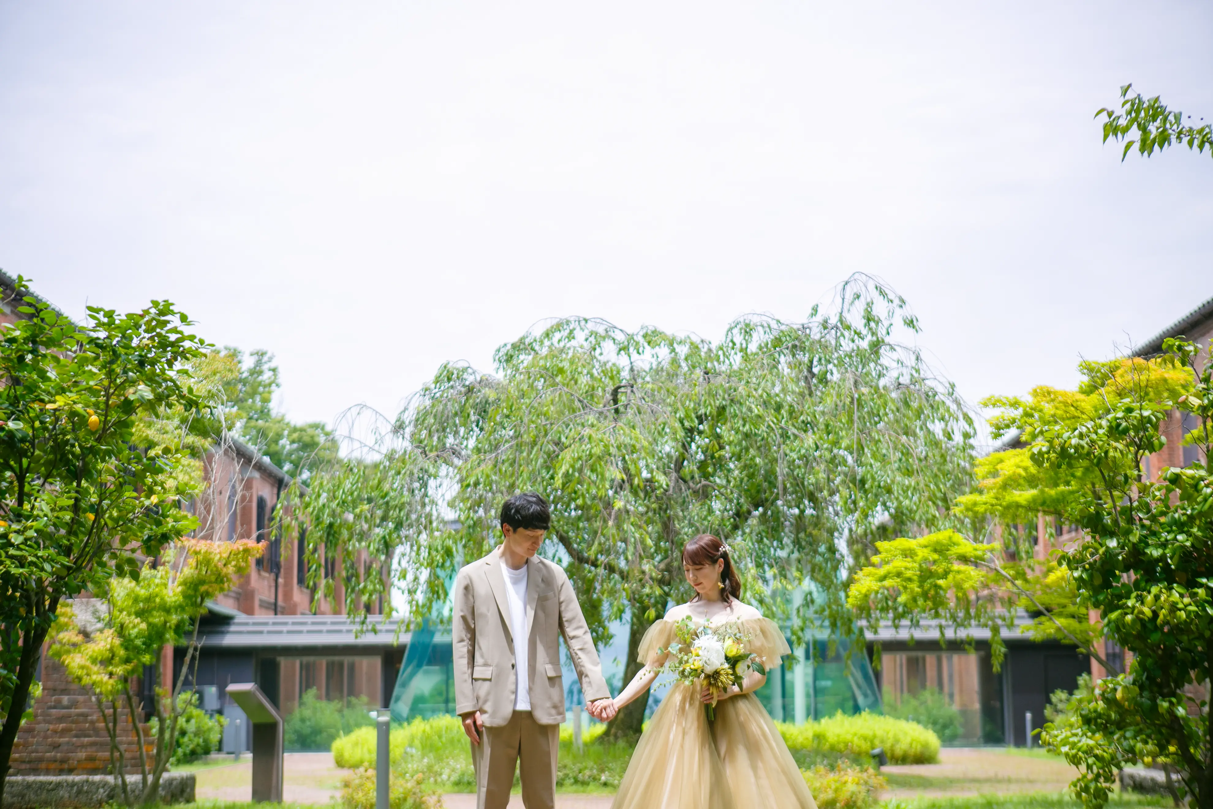 赤れんがミュージアムの中庭でしだれ桜の新緑と広い空を背景に手をつなぐ新郎新婦