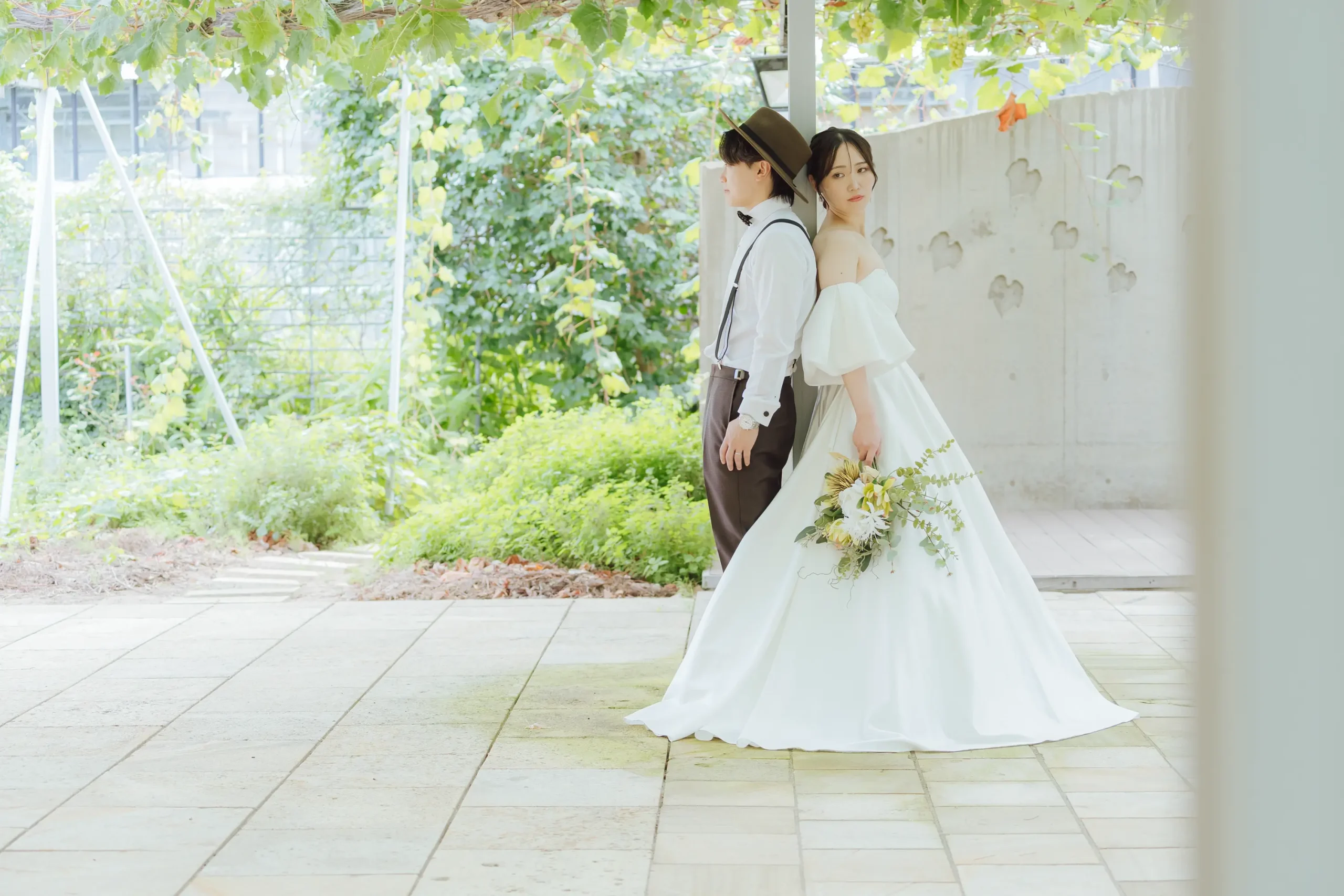 金沢・ぶどうの森のぶどう棚のあるテラスで背中合わせに立つ帽子の新郎とドレスの新婦