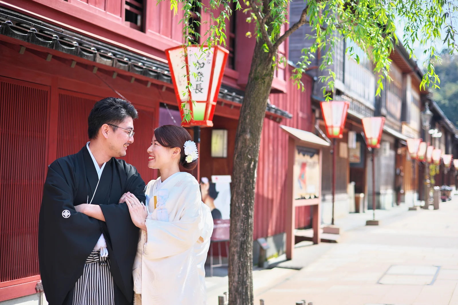 金沢・ひがし茶屋街の朱色の茶屋の前で向き合って微笑む白無垢と紋付袴の新郎新婦