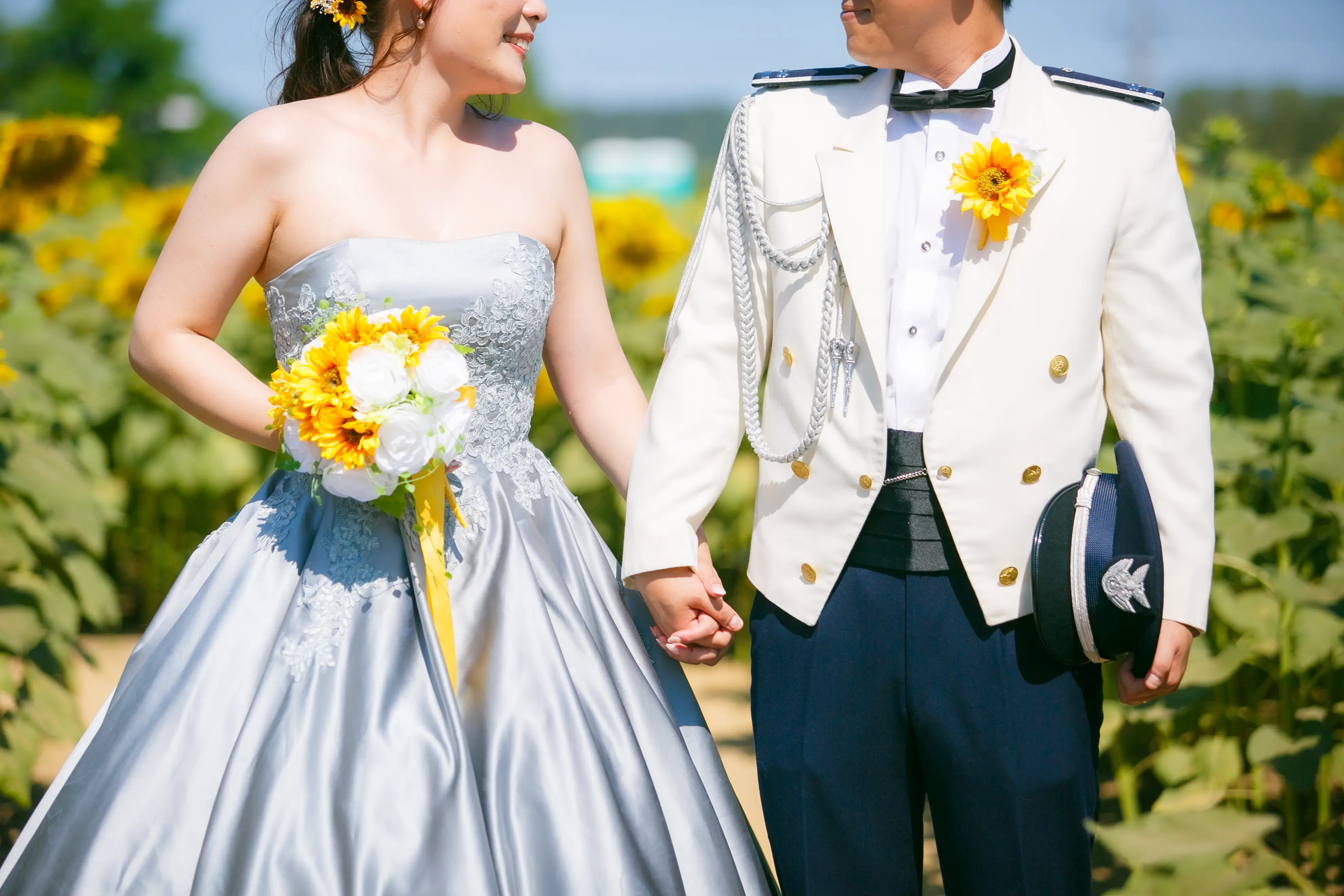 手をつなぐ新郎新婦の上半身。水色ドレスの新婦が持つひまわりのブーケと、ひまわりを挿した白い制服の新郎