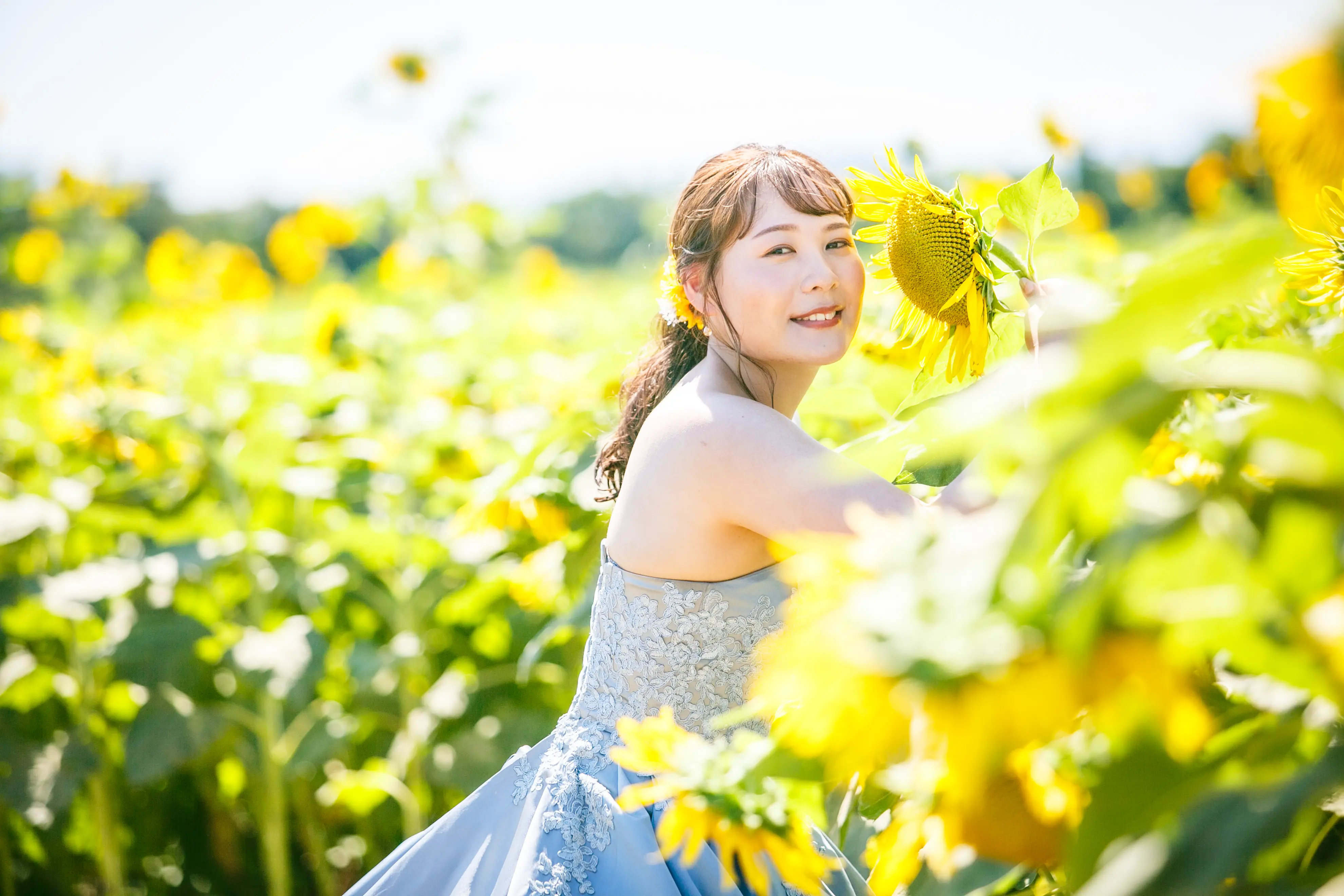 ひまわりの花に寄り添い振り返る、水色ドレスの新婦とひまわり畑
