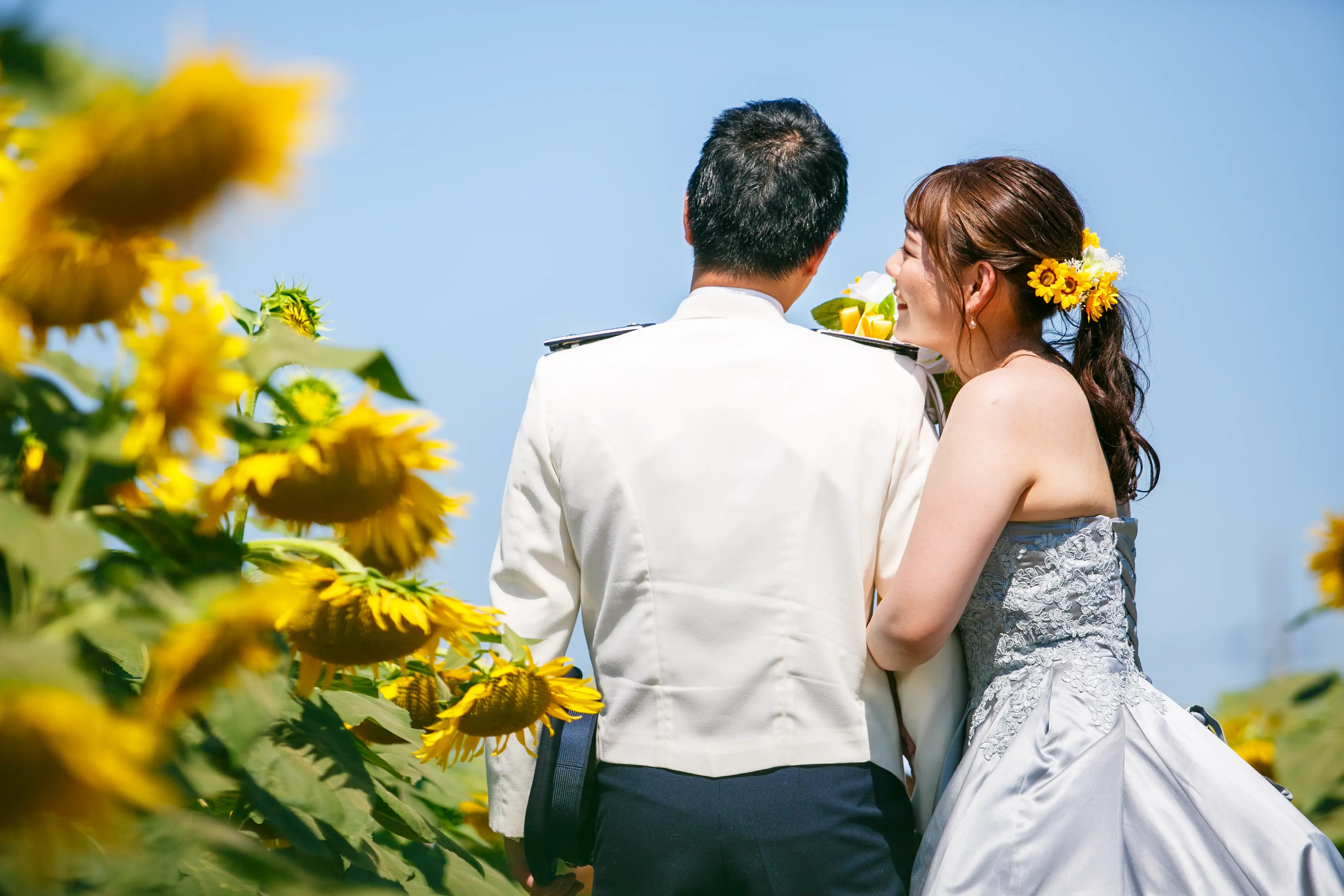 ひまわり畑と青空の中、背を向けて顔を寄せ合い笑う水色ドレスの新婦と白い制服姿の新郎