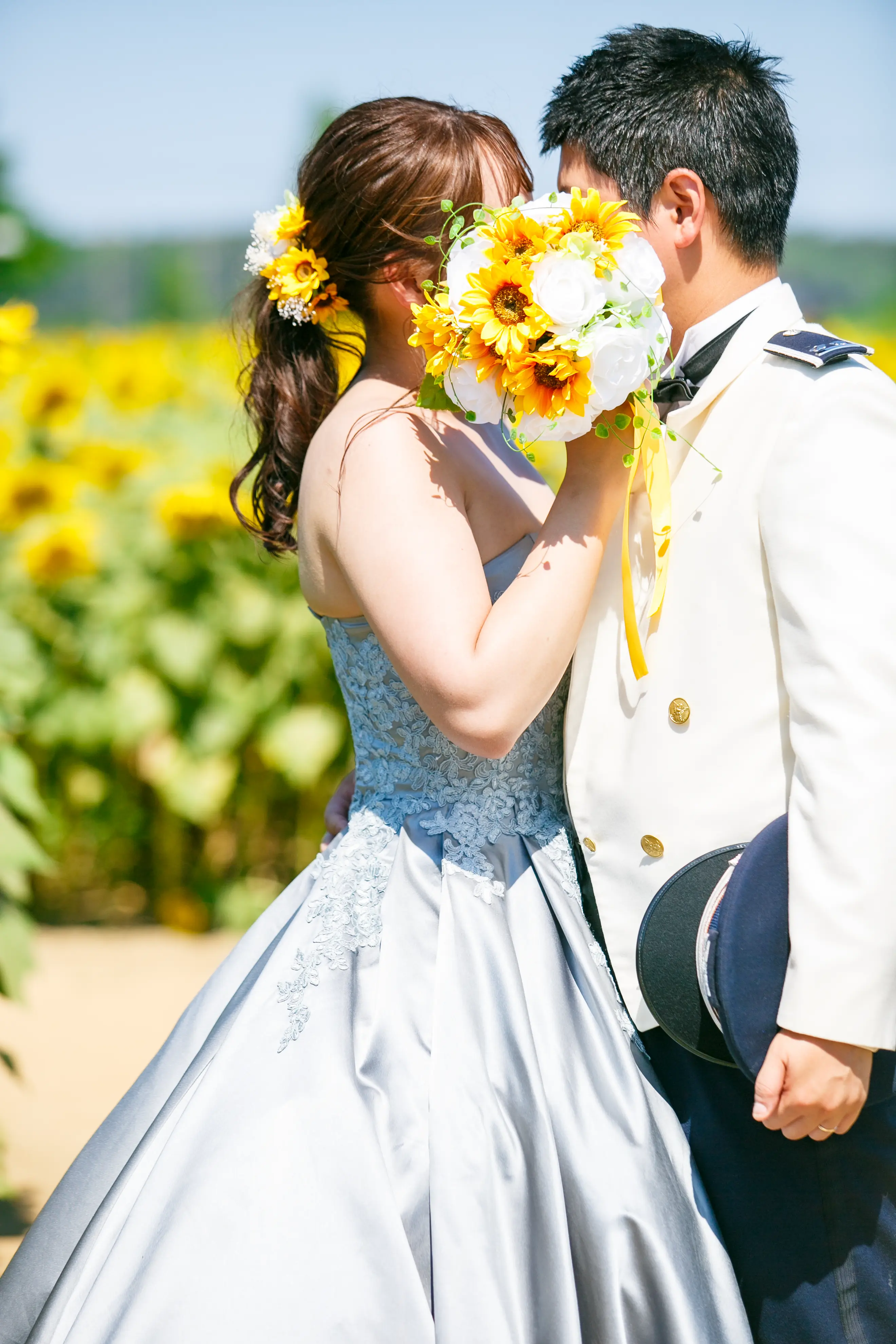 ひまわりのブーケごしに顔を寄せ合う、水色ドレスの新婦と白い制服姿の新郎