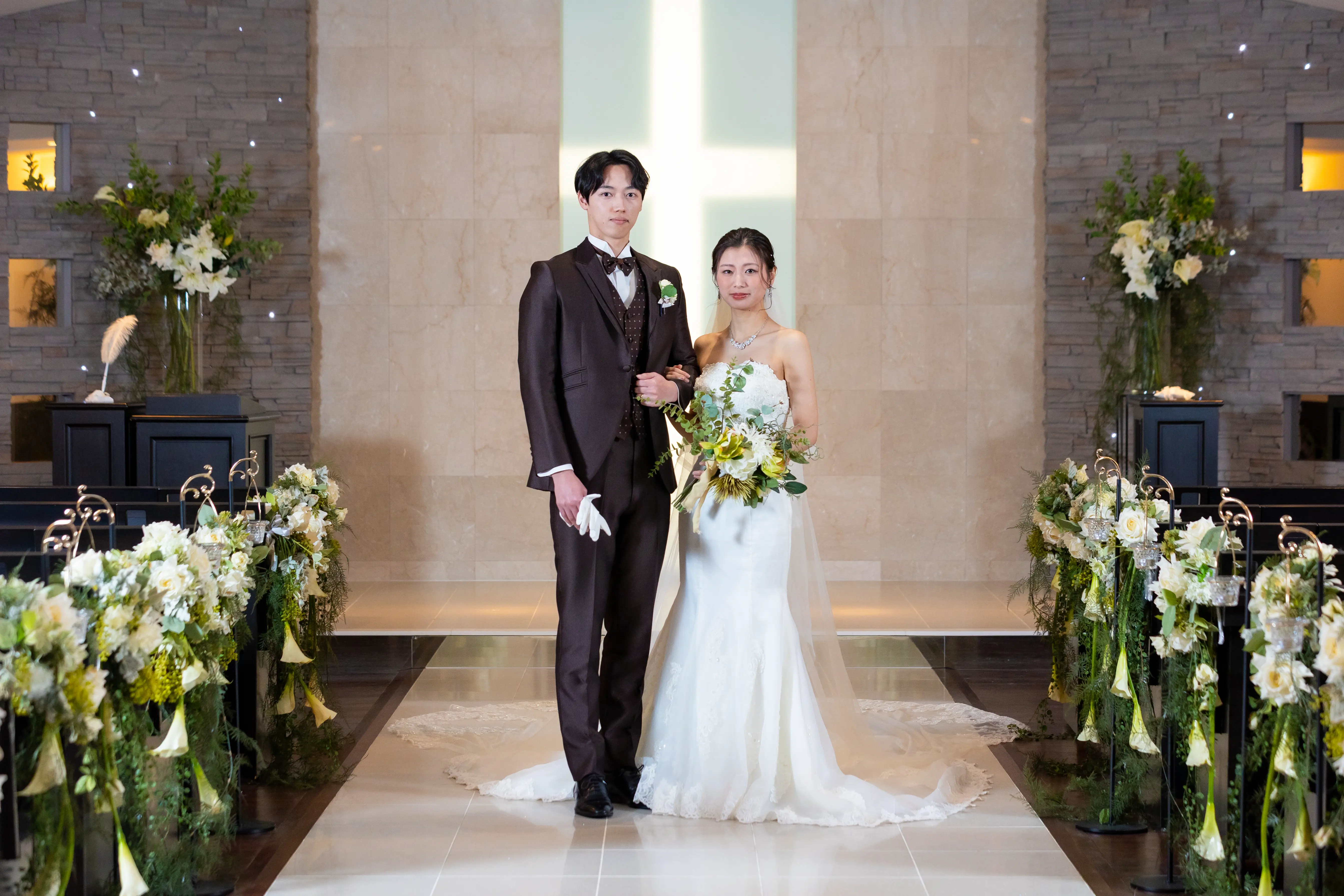 ホテル金沢のチャペル「アンジェラス」で祭壇前に並ぶウェディングドレス姿の新婦とブラウンのタキシード姿の新郎。背景に光る十字架と白い花のバージンロード装飾。