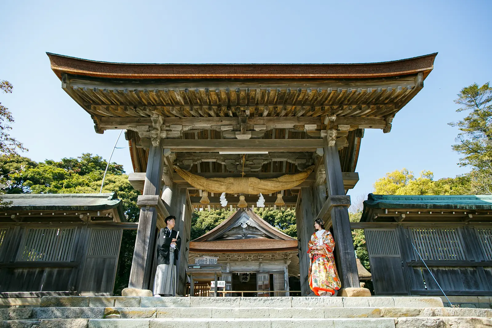 気多大社の壮大な神門を見上げる構図で両脇に立つ色打掛の新婦と紋付袴の新郎の和装フォトウェディング