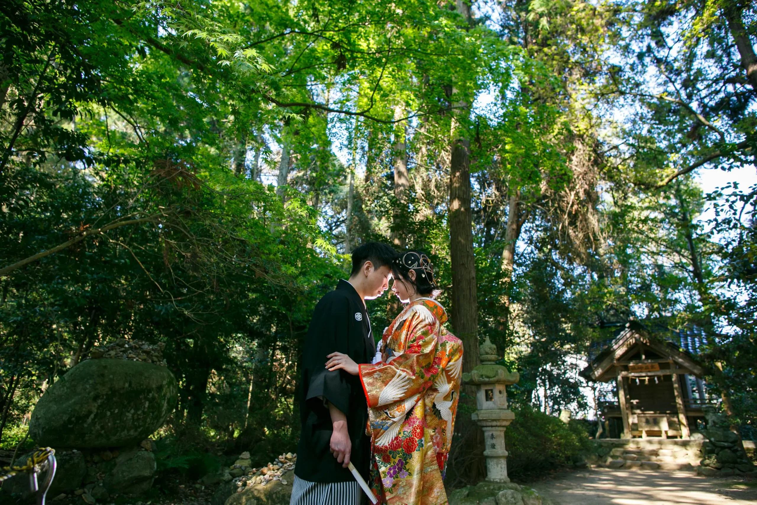 気多大社の緑の社叢と石灯籠を背景に額を寄せ合う鶴柄の色打掛の新婦と黒紋付袴の新郎
