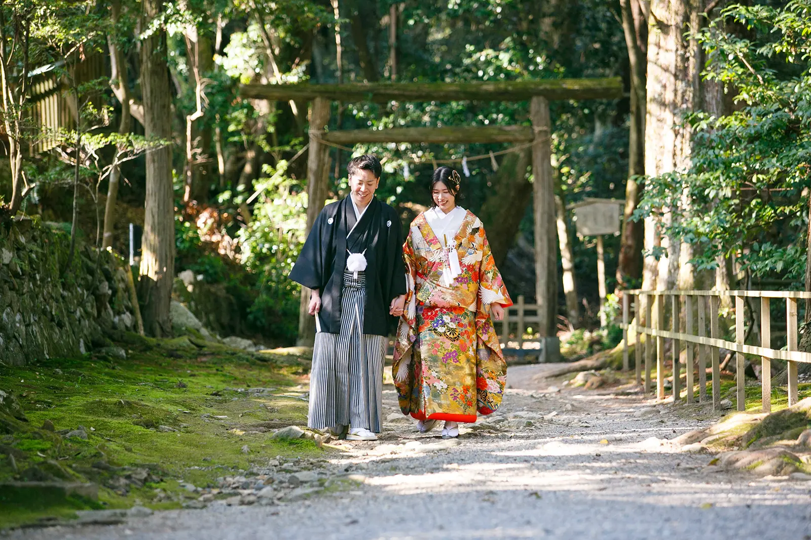 気多大社の木漏れ日射す参道と鳥居を背に手をつないで歩く色打掛の新婦と紋付袴の新郎