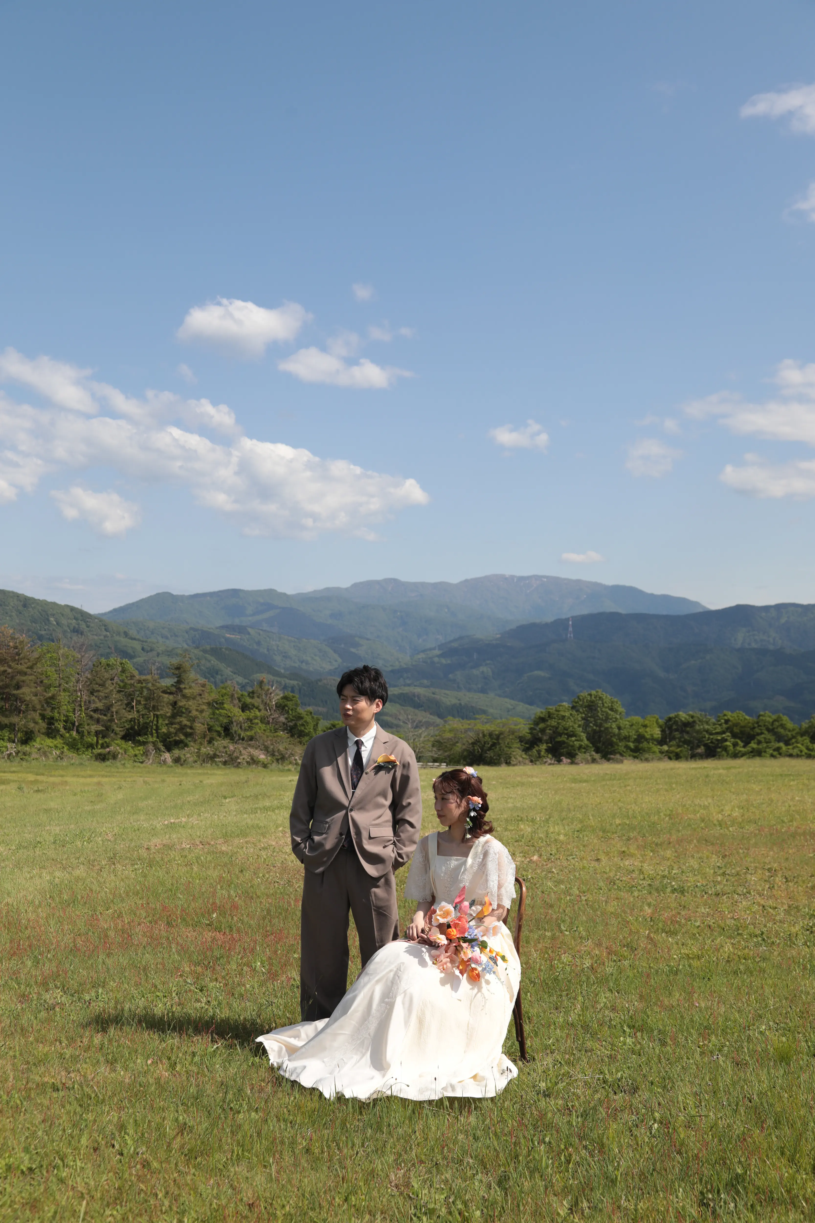 金沢のキゴ山で連なる新緑の山並みを背景に椅子に座りブーケを持つ洋装の新婦と隣に立つ新郎