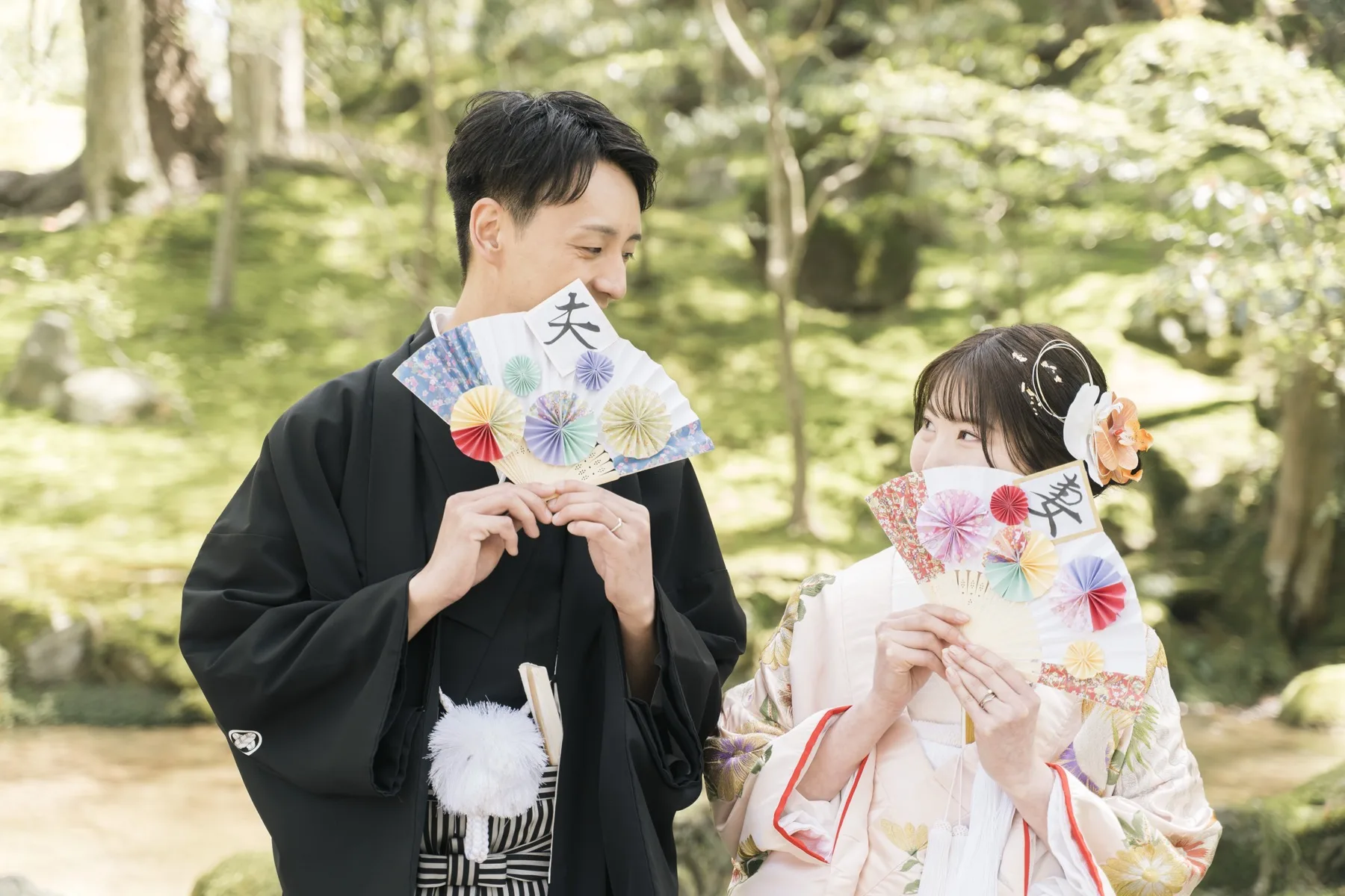 新緑の日本庭園で夫と婦の和柄の扇を手に見つめ合う、黒紋付の新郎と色打掛の花嫁｜金沢の和装ロケーション