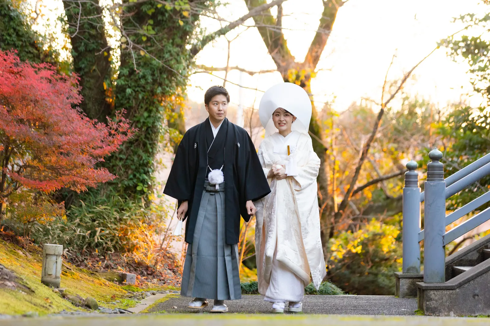 金沢国際ホテルの神殿の外、左手の紅葉と右手の橋の欄干を入れて引きで写した白無垢と紋付袴のふたり