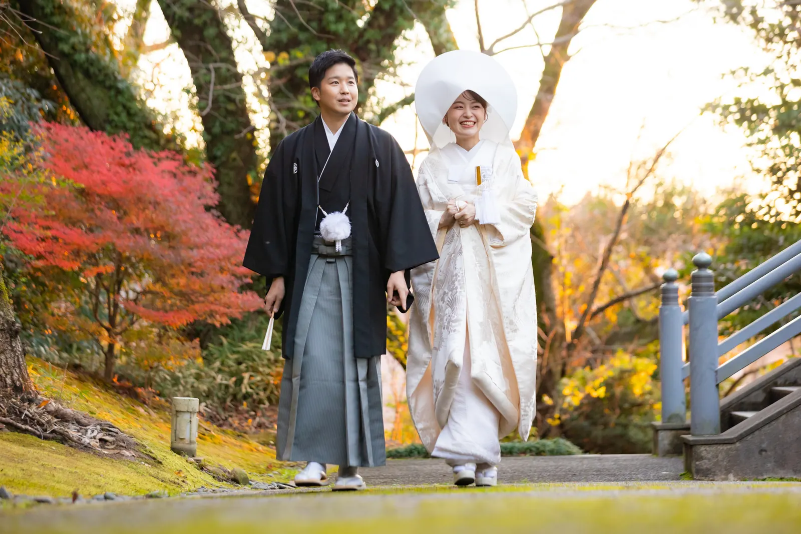金沢国際ホテルの神殿の外、紅葉を背景に歩きながら微笑み合う綿帽子の白無垢の花嫁と紋付袴の新郎
