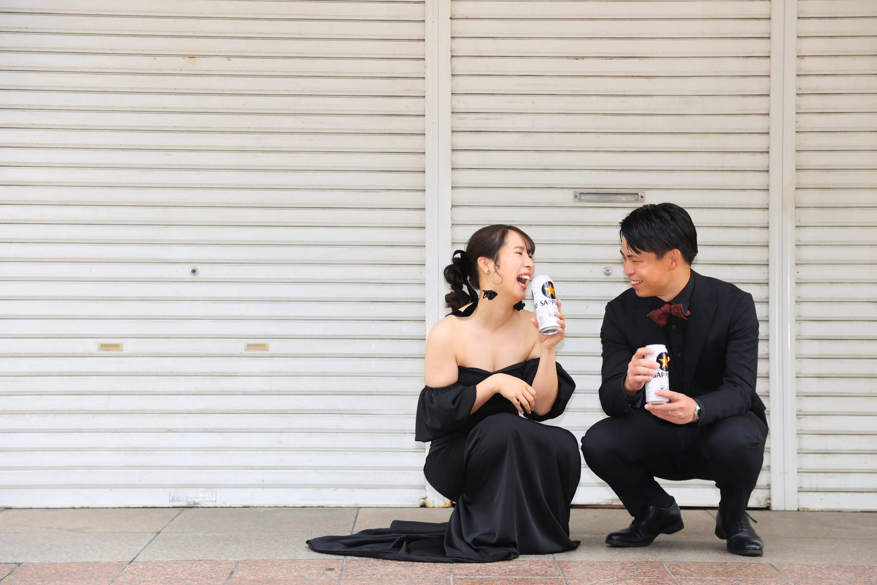 金沢の街の白いシャッターの前に座り缶を手に笑い合う黒いドレスの新婦と黒いスーツの新郎
