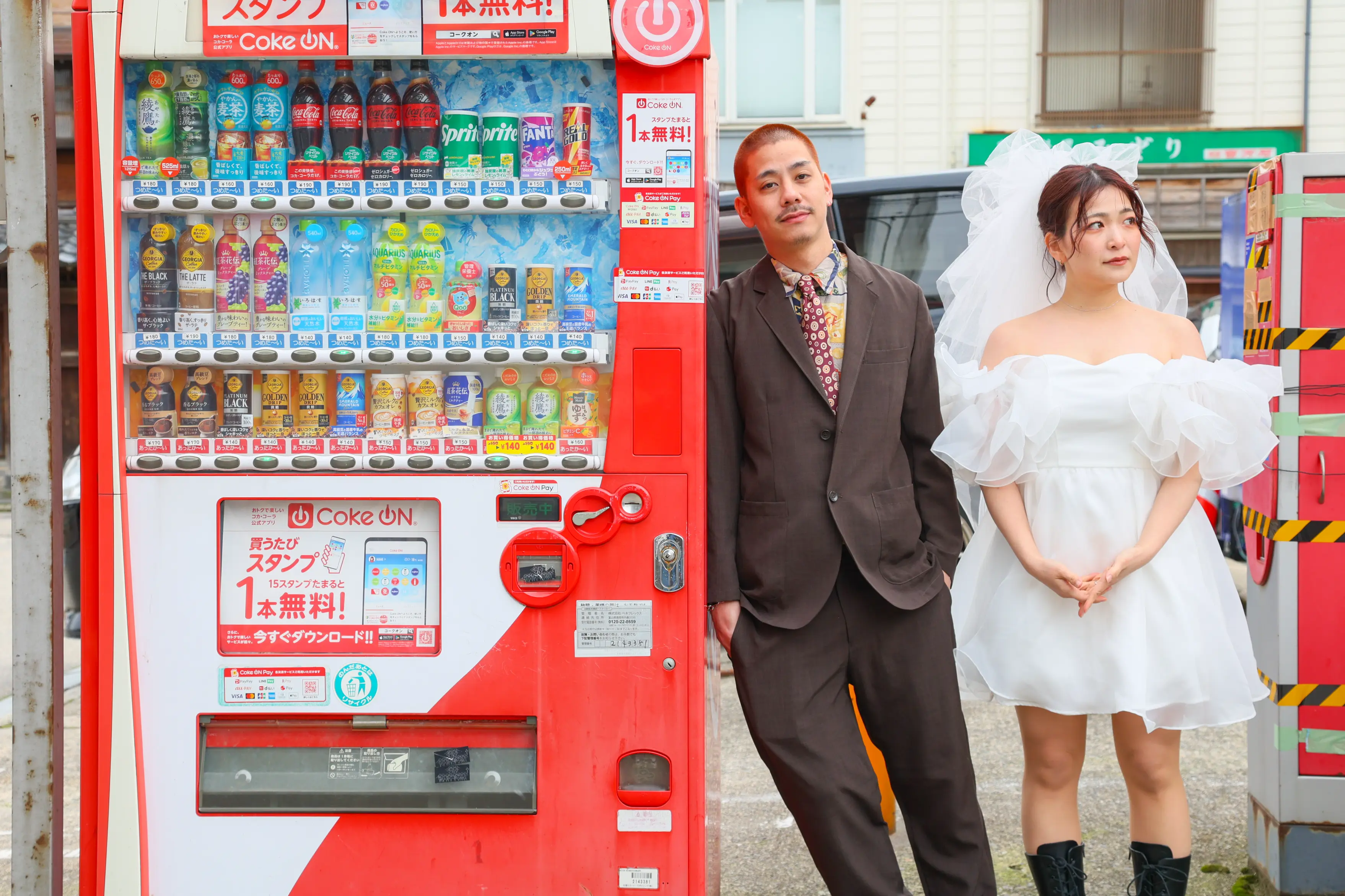 金沢の街角でコカ・コーラの自動販売機に寄りかかる白いミニドレスの新婦とブラウンのスーツの新郎
