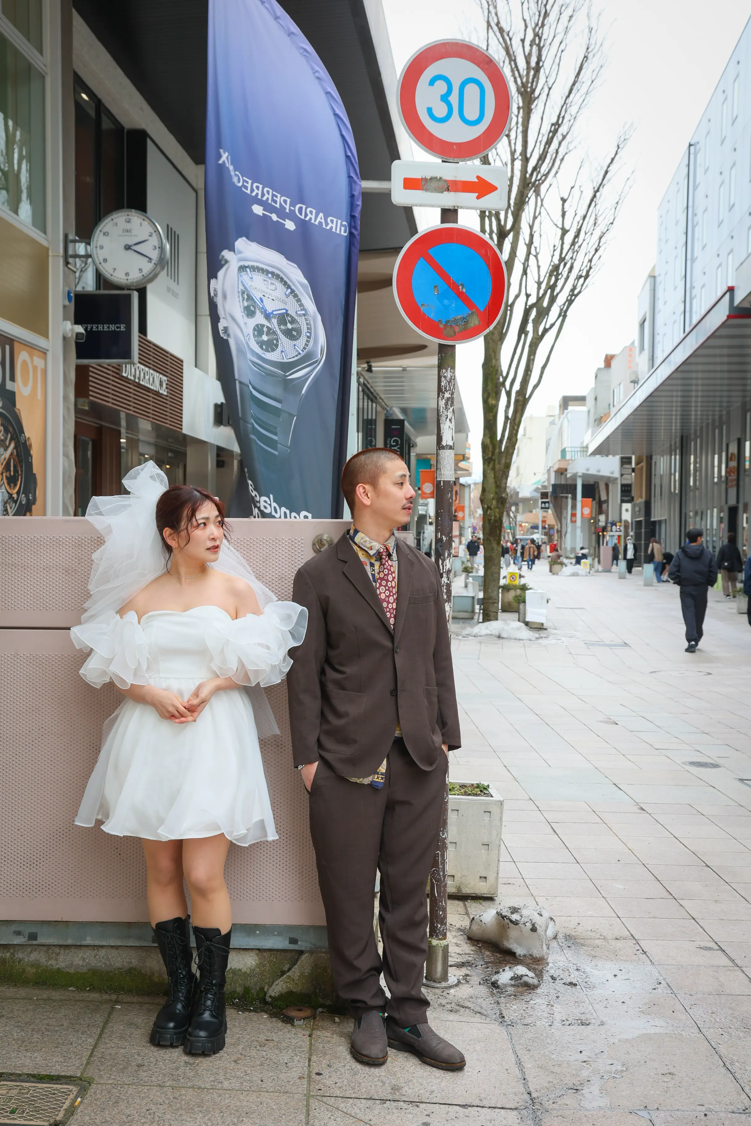 金沢・竪町の時計店が並ぶ通りで街灯の下に立つ白いミニドレスに黒いブーツの新婦とブラウンのスーツの新郎